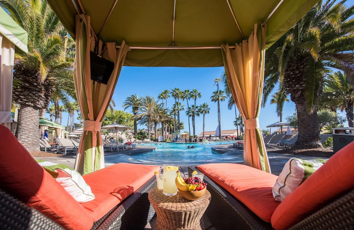A cozy cabana overlooking the pool at the vegan-friendly Mission Bay Resort in San Diego, California, with vibrant orange cushions, a refreshing fruit platter, and palm trees in the background.