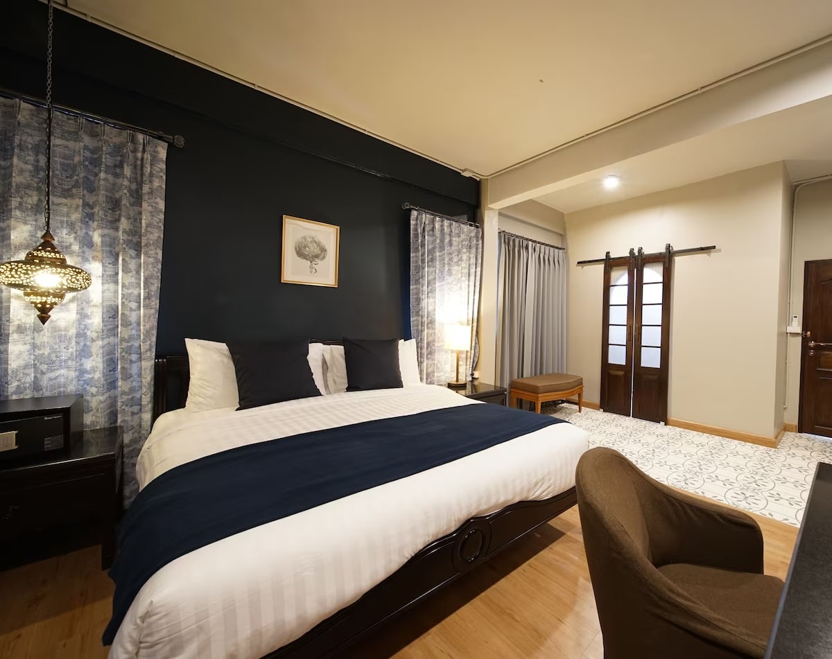 A cozy and elegantly decorated hotel room at Na Vayla PaPlern Hotel in Bangkok, Thailand. The room features a large bed with white linens and dark blue accents, a stylish hanging lamp, and framed art on a dark accent wall. There are sliding wooden doors, a small bench, and a comfortable chair next to a desk, creating a warm and inviting atmosphere.