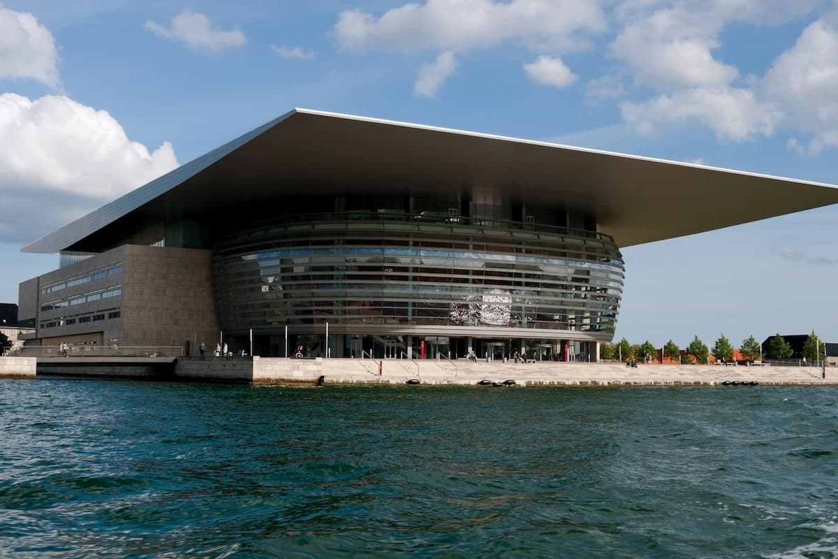 The Copenhagen Opera House, a modern architectural landmark in Denmark, situated along the waterfront with its striking curved glass facade and expansive overhanging roof.
