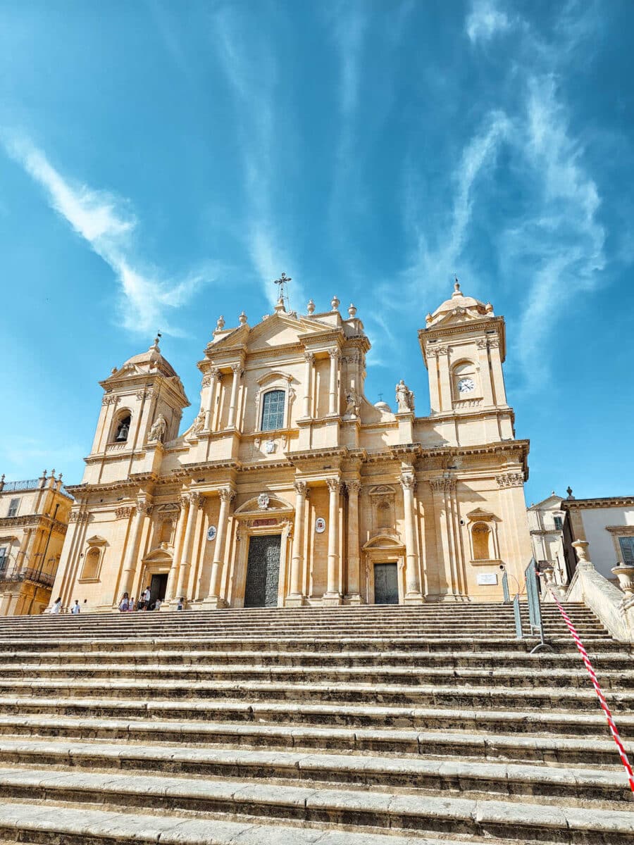 A beautiful view of the Noto Cathedral's stunning baroque façade in Sicily. The sun casts a warm glow on the light-colored limestone, highlighting the intricate architectural details and the grand staircase leading up to the entrance.