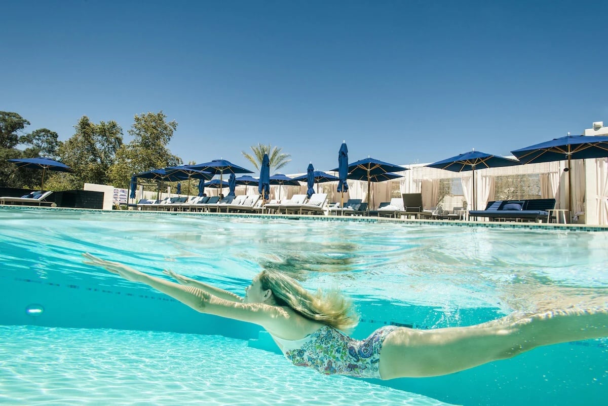 A guest swimming in the sparkling outdoor pool at the vegan-friendly Ojai Valley Inn in Ojai, California, surrounded by lounge chairs and blue umbrellas under a clear sky.