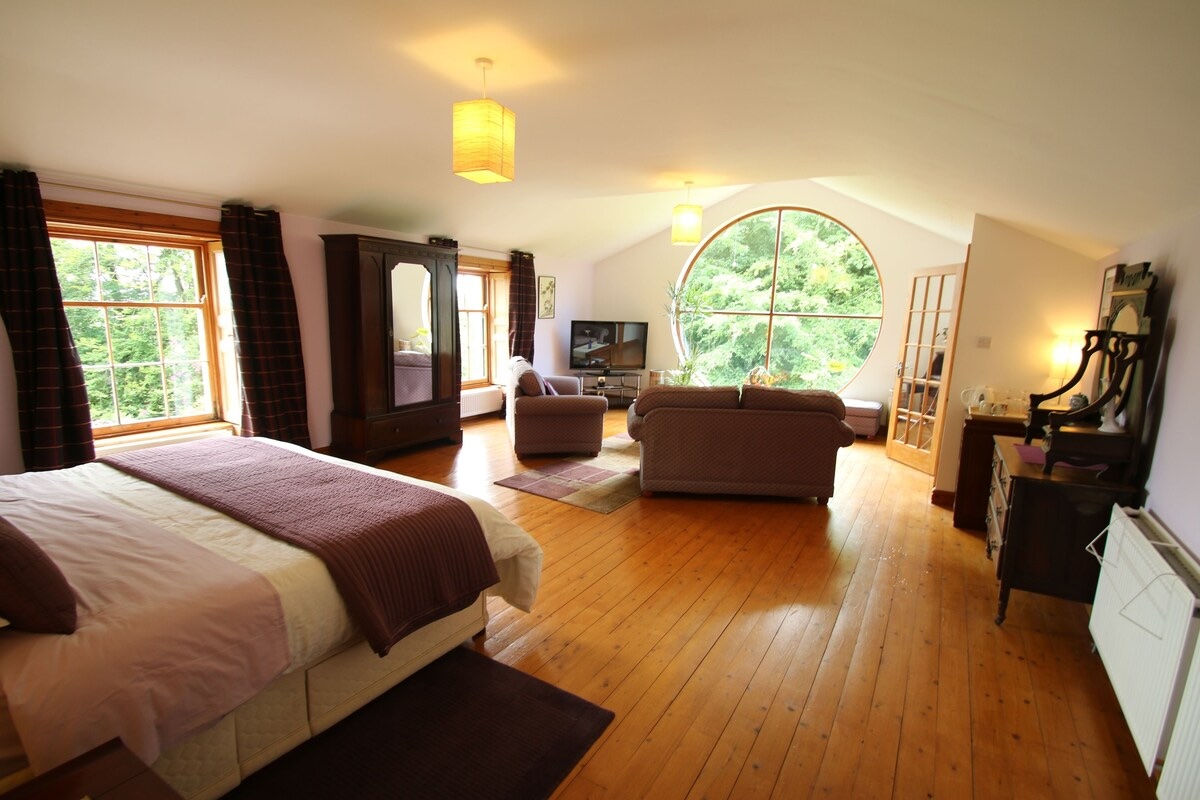 A spacious and bright bedroom at the Over the Rainbow - Vegetarian Guesthouse in Wales with a large bed, wooden furniture, and a cozy seating area. The room features a circular window with a view of lush greenery, wooden floors, and soft lighting from hanging lamps.
