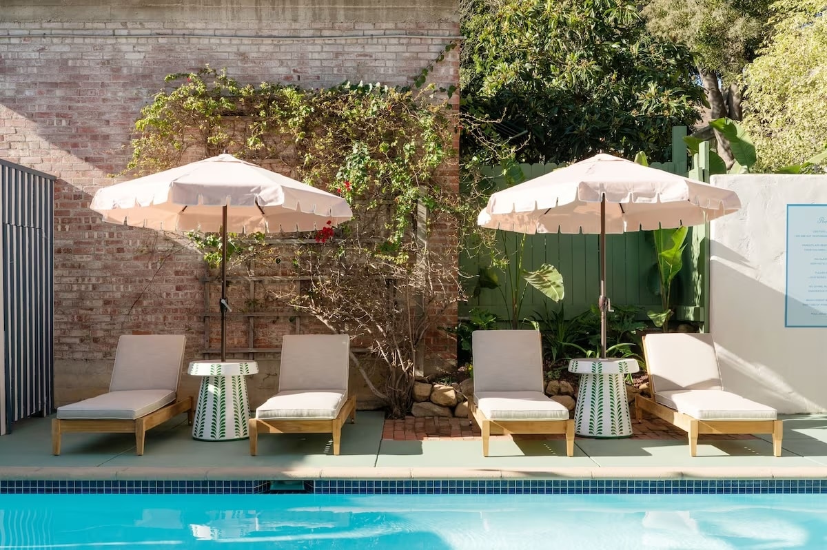 A poolside area at Palihouse Santa Barbara in Santa Barbara, California, featuring stylish lounge chairs, pastel umbrellas, and lush greenery against a brick wall backdrop.