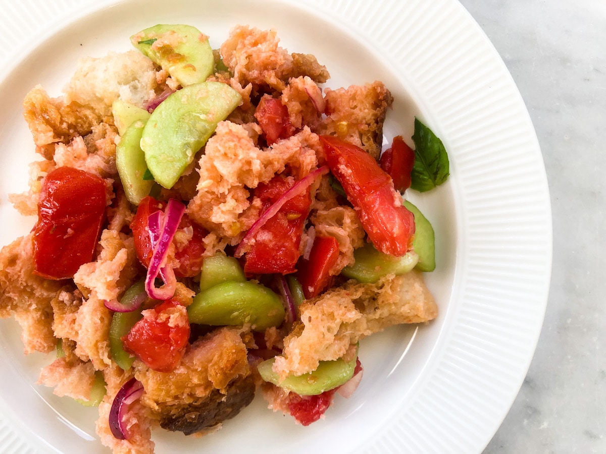 Panzanella salad made with chunks of soaked stale bread, sliced cucumbers, tomatoes, red onions, and fresh basil, served on a white plate.