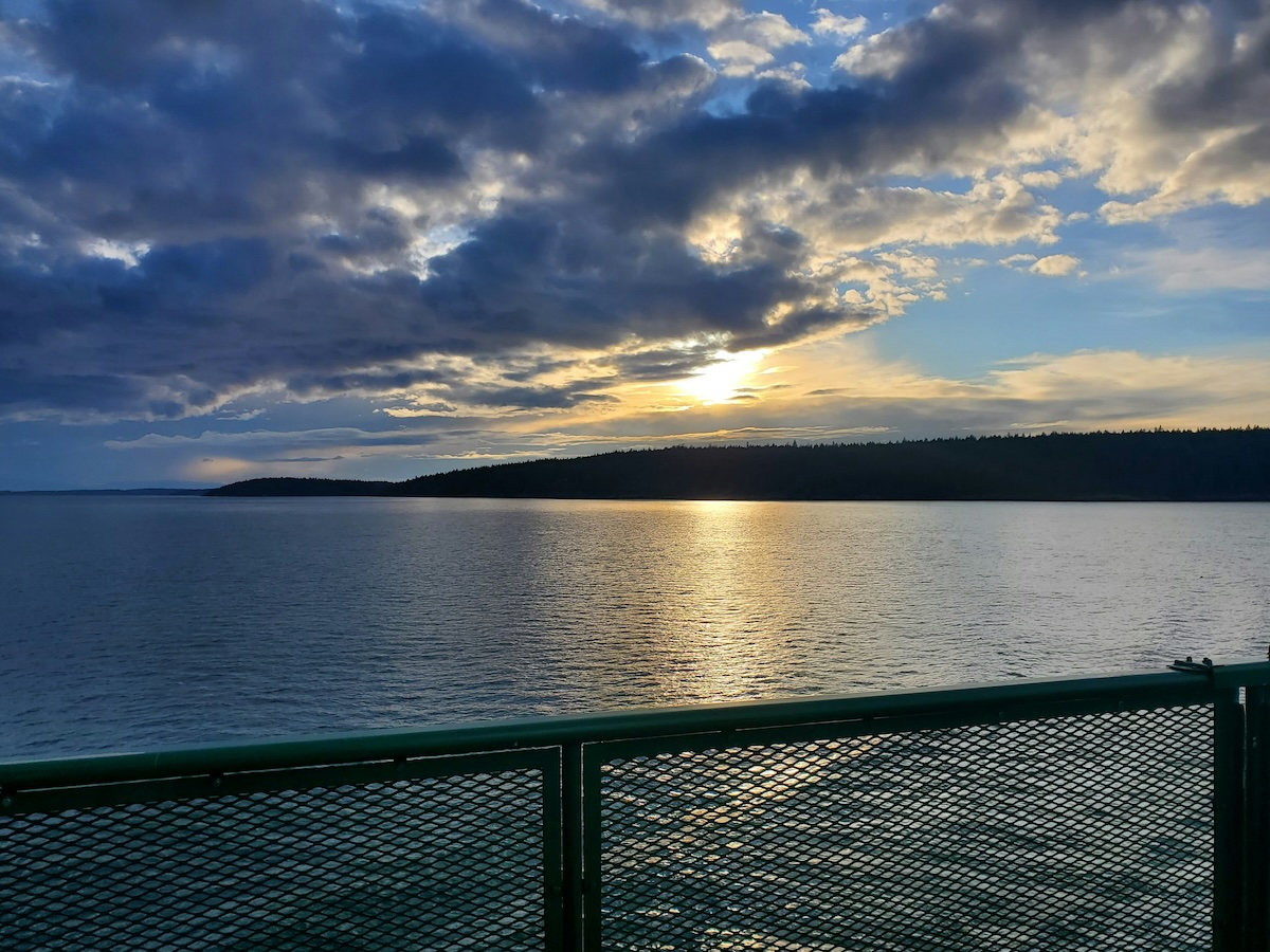 Stunning sunset view over the water from a ferry at Eastsound, Orcas Island, Washington. The sun dips below the horizon, casting a golden reflection on the calm waters, with clouds adding dramatic contrast to the sky.