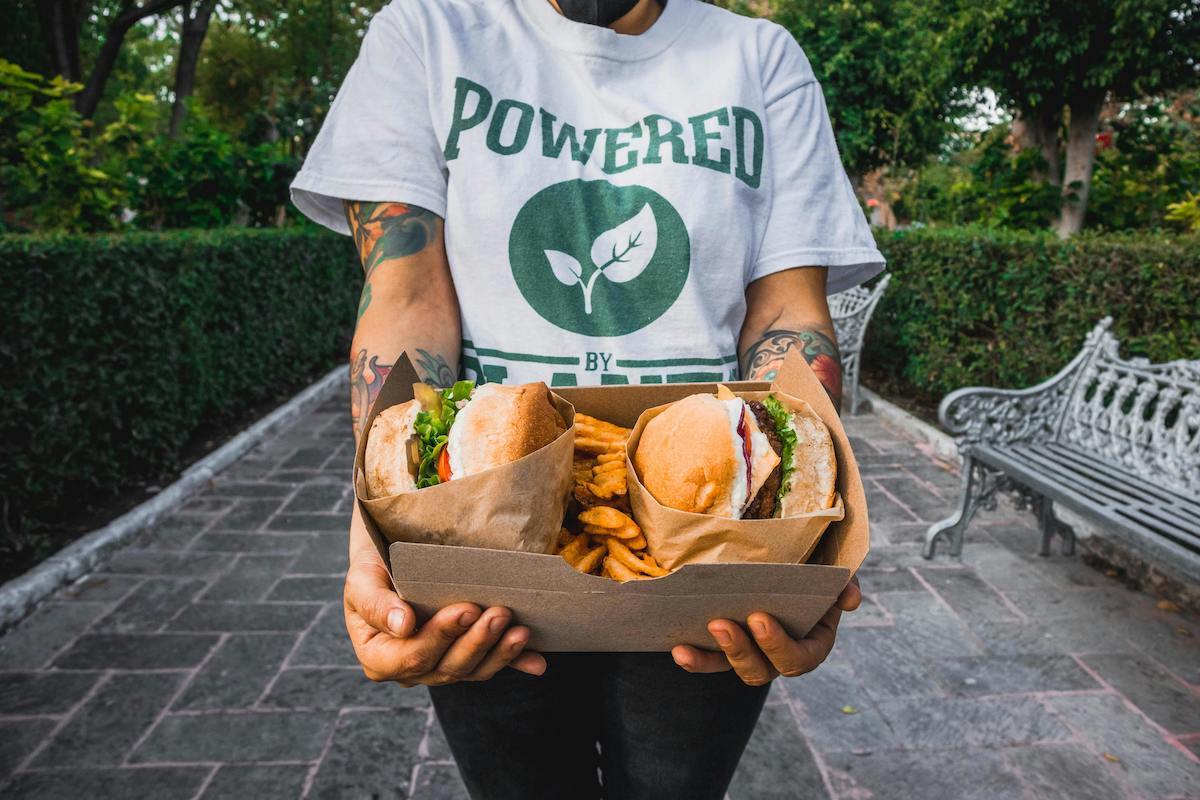 A person wearing a white shirt that says "Powered by Plants" while holding a cardboard tray containing two vegan burgers wrapped in paper, accompanied by a serving of crinkle-cut fries.