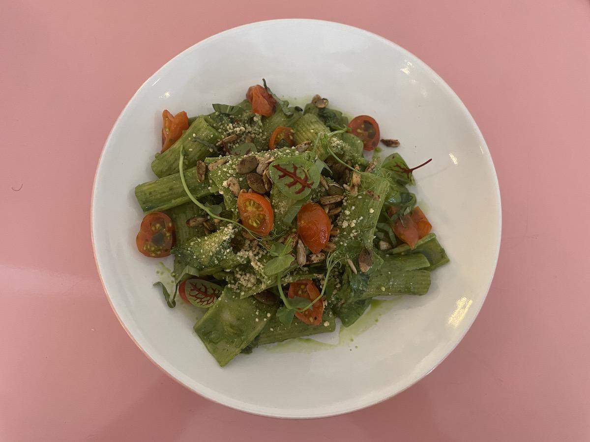 A dish of pesto pasta served in a white bowl against a pink background. The pasta is coated in green pesto sauce, garnished with cherry tomatoes, leafy greens, and sprinkled with seeds and a light dusting of vegan cheese or nuts