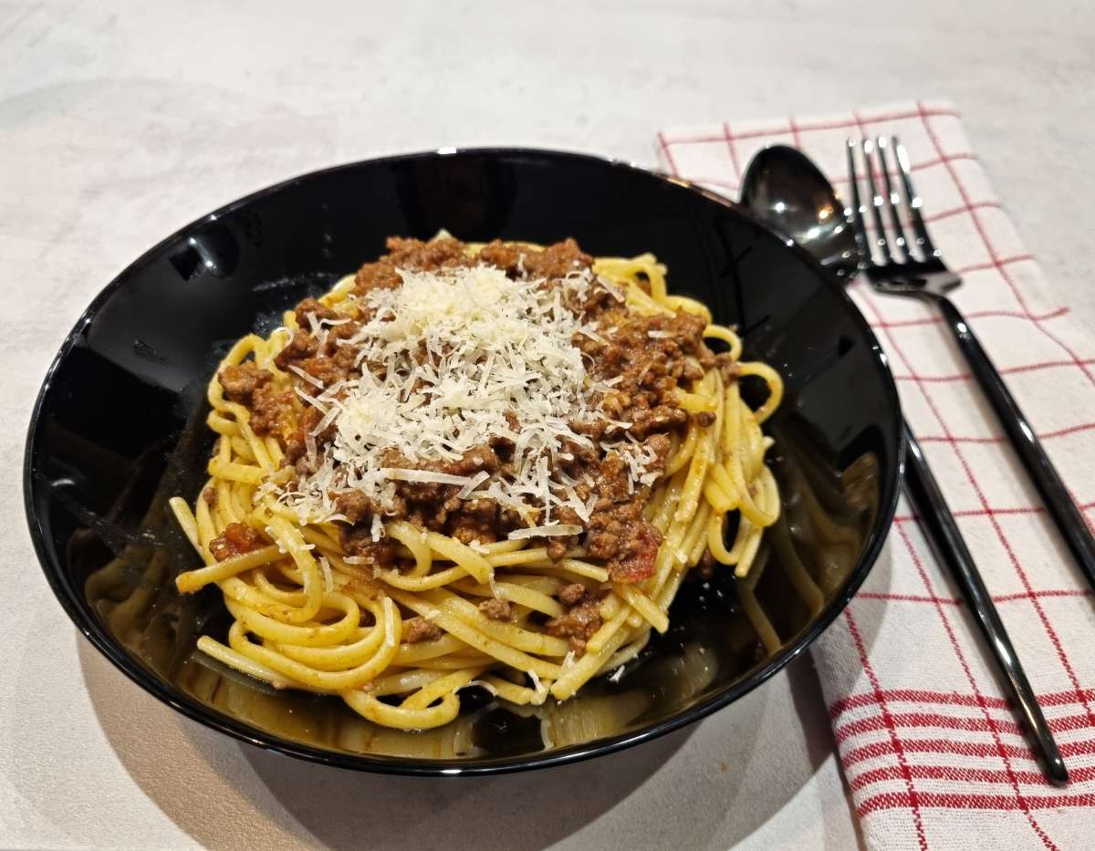 A bowl of tagliatelle pasta topped with homemade ragu Bolognese and freshly grated Parmigiano Reggiano, served in a black dish with utensils on a red-and-white checkered napkin.