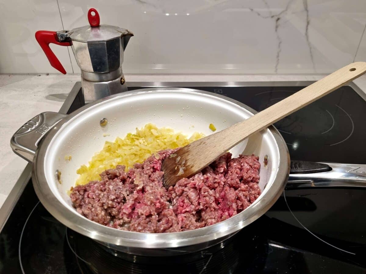 Raw ground beef and sautéed onions cooking in a stainless steel pan, stirred with a wooden spoon, with a stovetop espresso maker visible in the background.