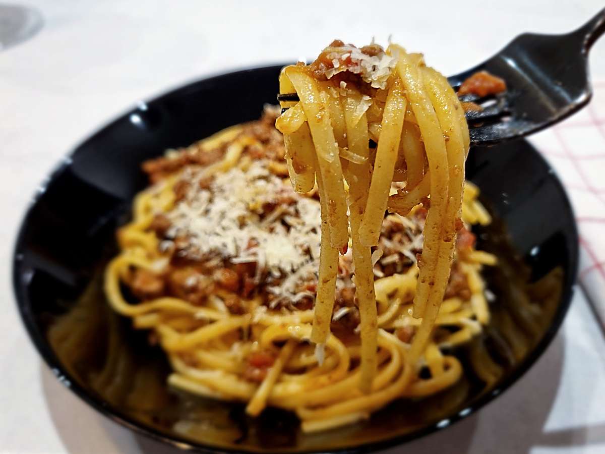 A close-up of tagliatelle pasta with homemade ragu Bolognese, topped with grated Parmigiano Reggiano. A fork lifts a twirl of saucy noodles from a black bowl.