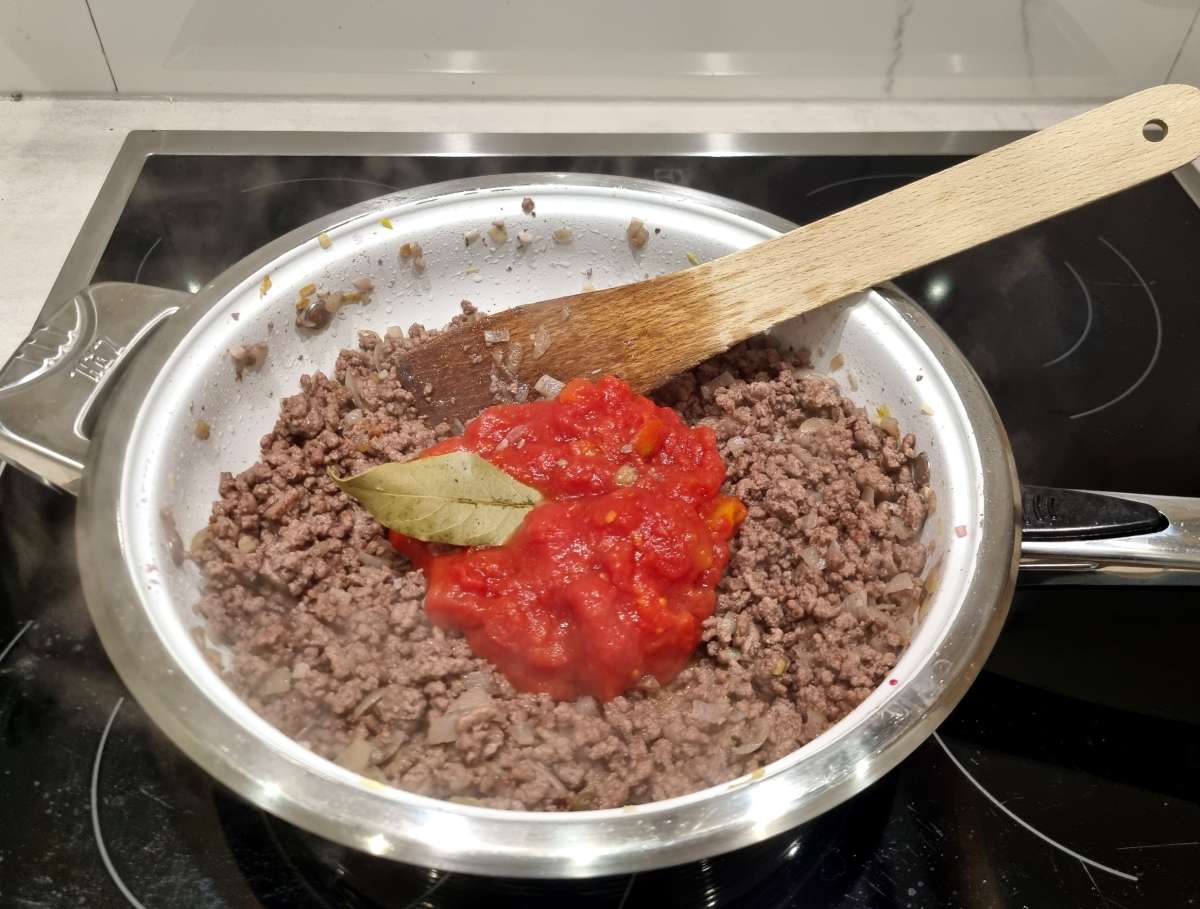 Ground beef cooking in a stainless steel pan with sautéed onions, topped with tomato sauce and a bay leaf, with a wooden spoon resting on the edge.