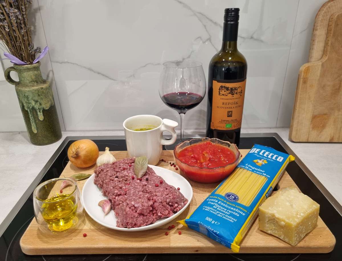 Ingredients for homemade ragu Bolognese laid out on a wooden board, including ground beef, garlic, onion, olive oil, tomato sauce, vegetable stock, linguine pasta, Parmigiano Reggiano, and a bottle of Refosk red wine with a filled glass.