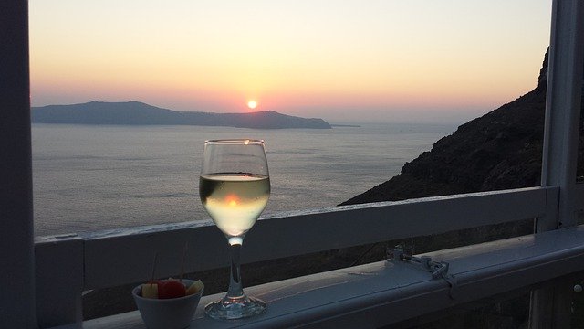 glass of white wine on a window sill with a view of the sun setting over rolling hills in Santorini, Greece