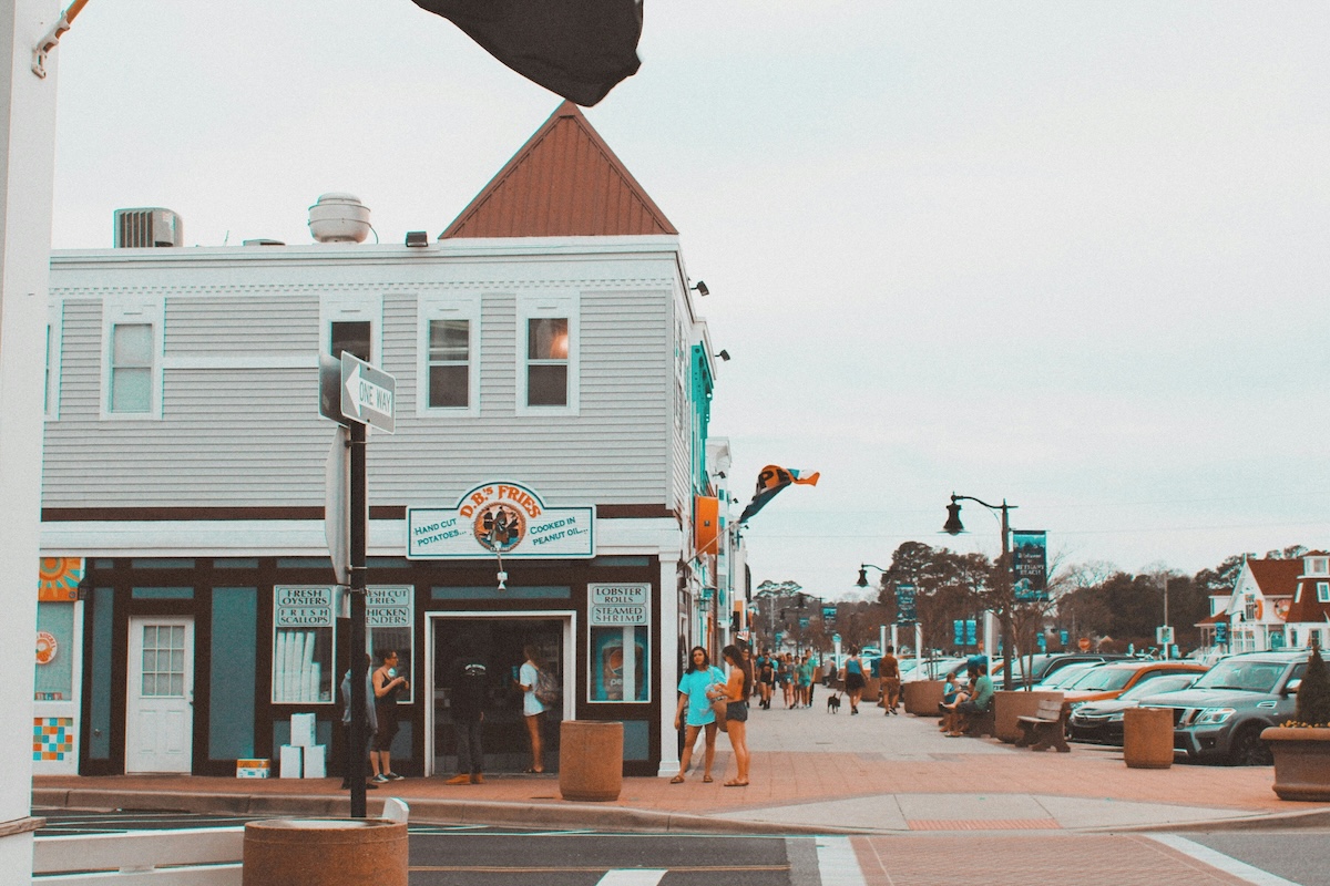 Street view of Bethany Beach, Delaware, featuring a quaint coastal town with a small restaurant and people walking along the sidewalk. The scene captures the relaxed, laid-back atmosphere of this beach destination.