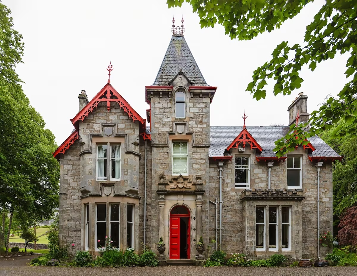 A charming stone building at the vegan Saorsa 1875 hotel in Scotland with Victorian architecture, featuring a slate roof and ornate red trim. The central tower is flanked by two gabled wings, each with large windows. The building has a vibrant red door and is surrounded by lush greenery, including mature trees and well-maintained shrubs and flowers.