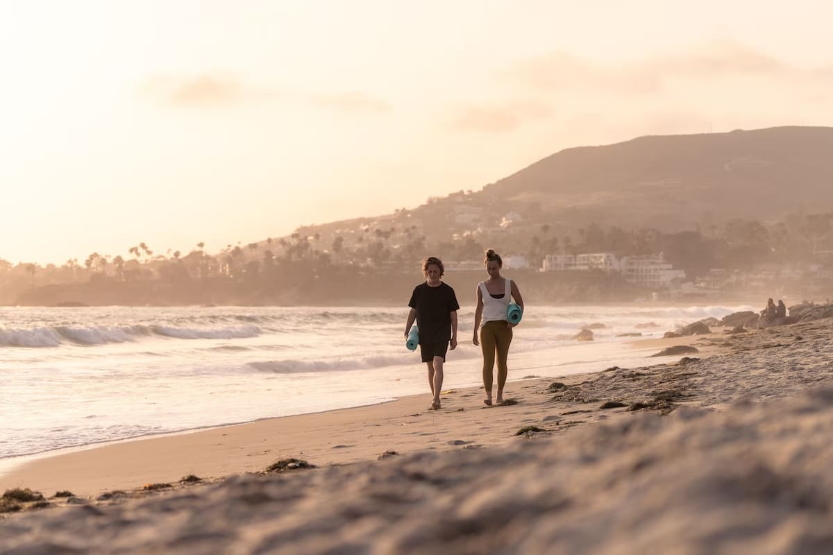 Two people walking along a serene beach at sunset near SCP Seven4One vegan hotel in Laguna Beach, California, with rolling waves and coastal hills in the background