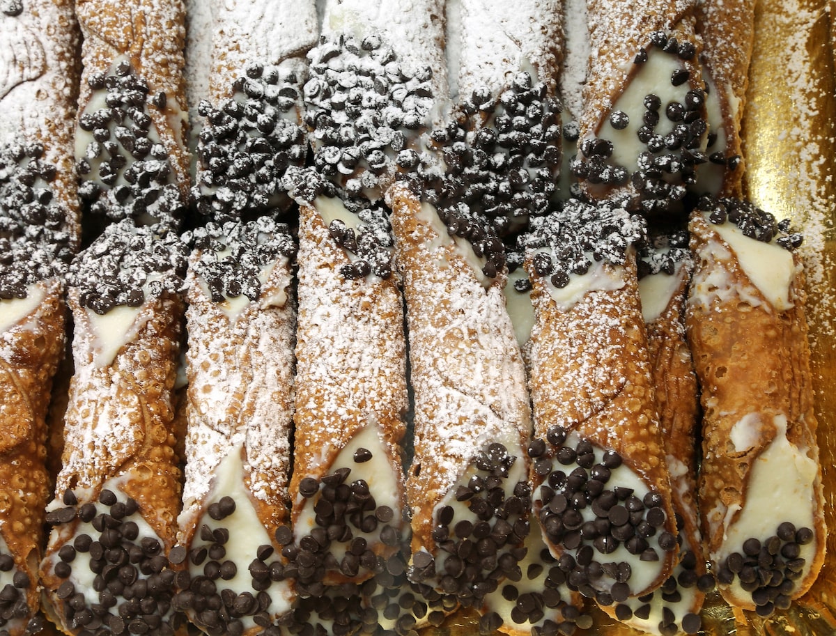 Rows of Sicilian cannoli filled with ricotta cream and topped with chocolate chips, displayed in a bakery setting.
