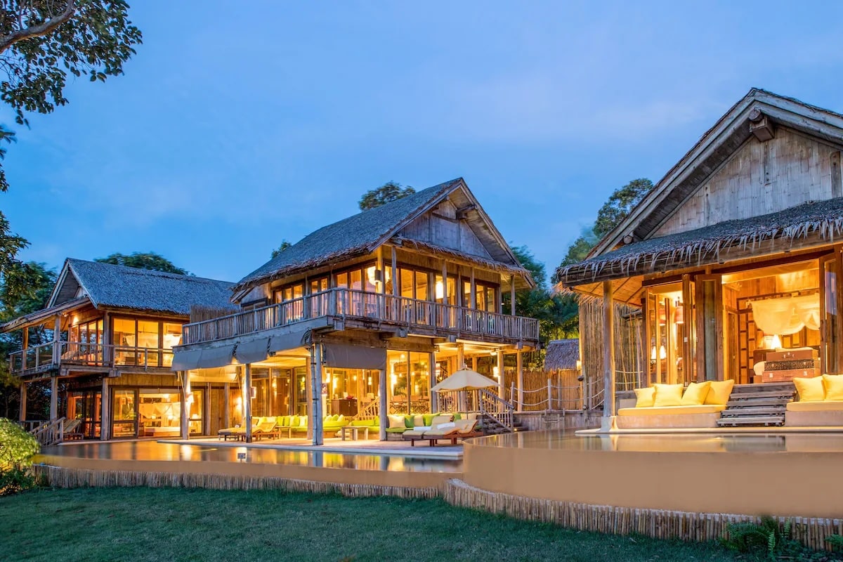 an illuminated tropical villa at the luxurious Soneva Kiri vegan resort in Thailand at dusk with thatched roofs and wooden structures. The villa features multiple stories and large glass windows, with a spacious outdoor area including lounge chairs and a pool. The surrounding area is lush with greenery, contributing to a serene and luxurious ambiance.