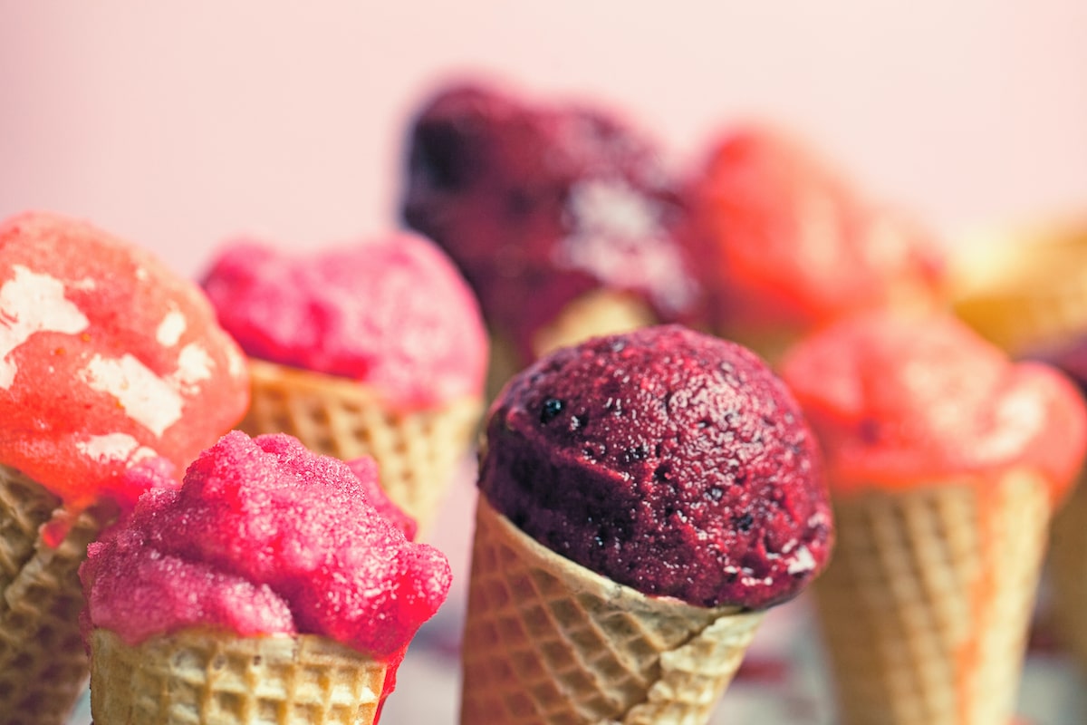 Assorted colorful fruit-flavored sorbetto scoops in waffle cones, including raspberry, blackberry, and citrus, displayed against a soft pink background.