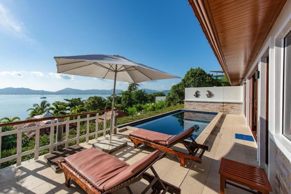 A stunning view from the terrace at Soul Villas by the Beach, a vegan friendly resort in Phuket, Thailand. The terrace features lounge chairs with cushions, an umbrella for shade, and a private pool. The scene overlooks a beautiful coastline with lush greenery and distant mountains, highlighting the serene and picturesque setting of this beach resort.