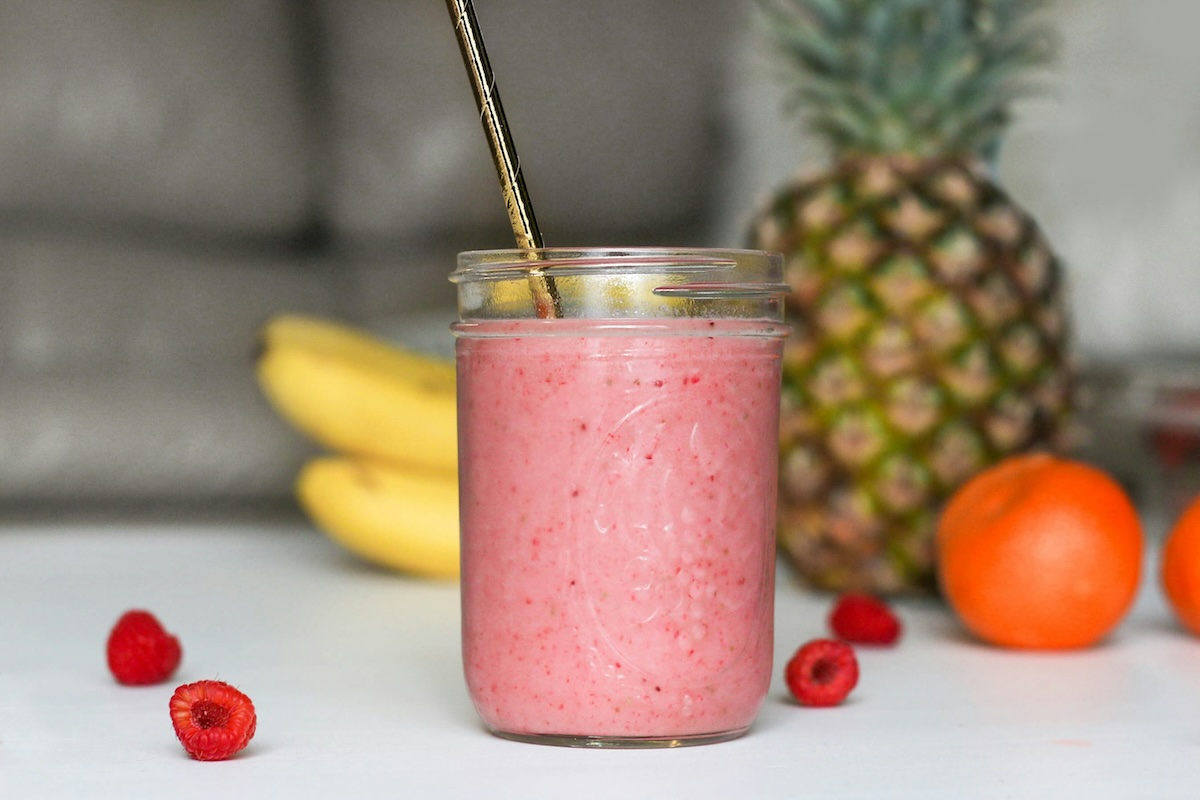 A vibrant pink smoothie in a mason jar with a metal straw, surrounded by fresh fruits including bananas, pineapple, and raspberries.