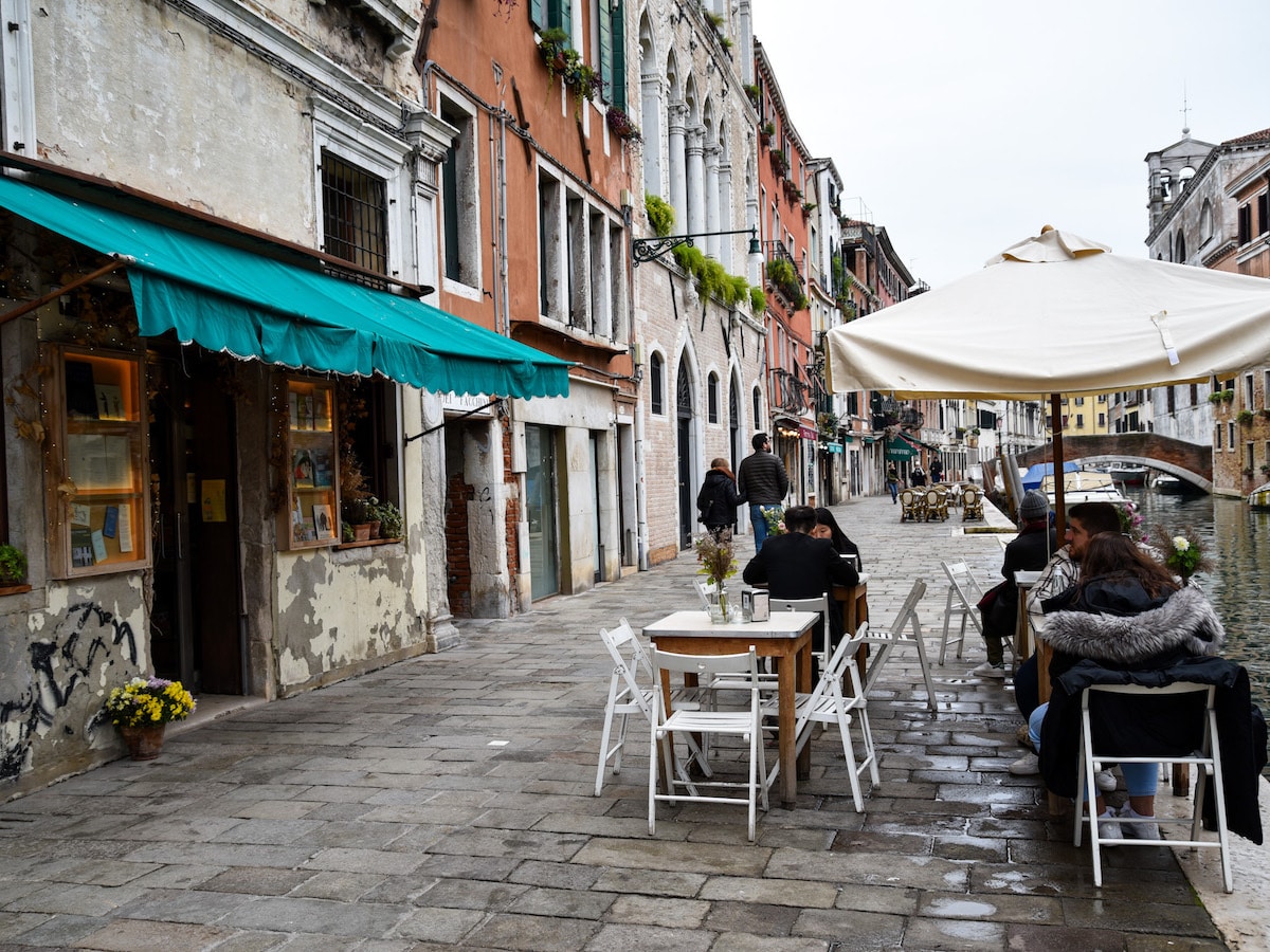 Outdoor canal-side seating at Sullaluna, a vegetarian and vegan café and bookshop in Venice, with white tables and chairs set under a large umbrella along a quiet cobblestone street.