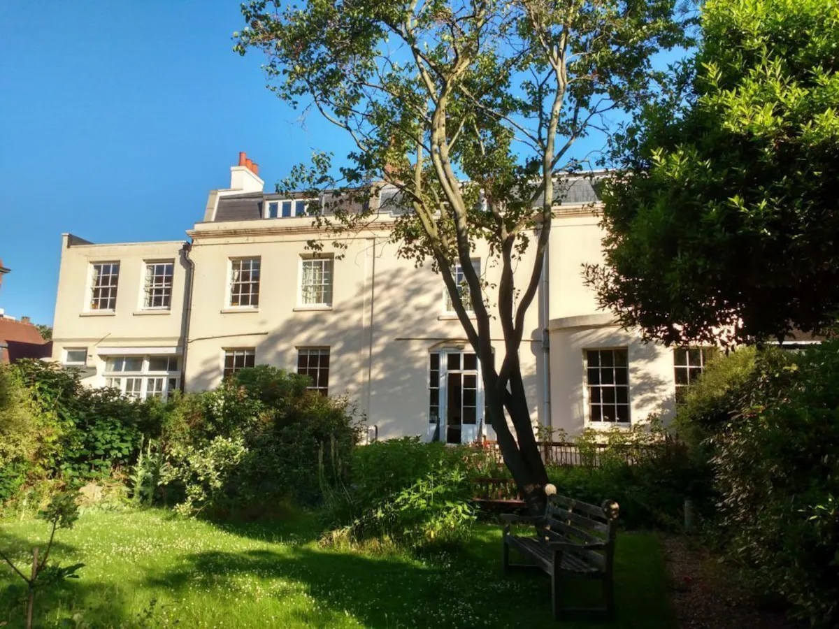 Elegant white building of Temple Lodge Club vegan hotel in London, featuring a lush garden and tall trees on a sunny day.