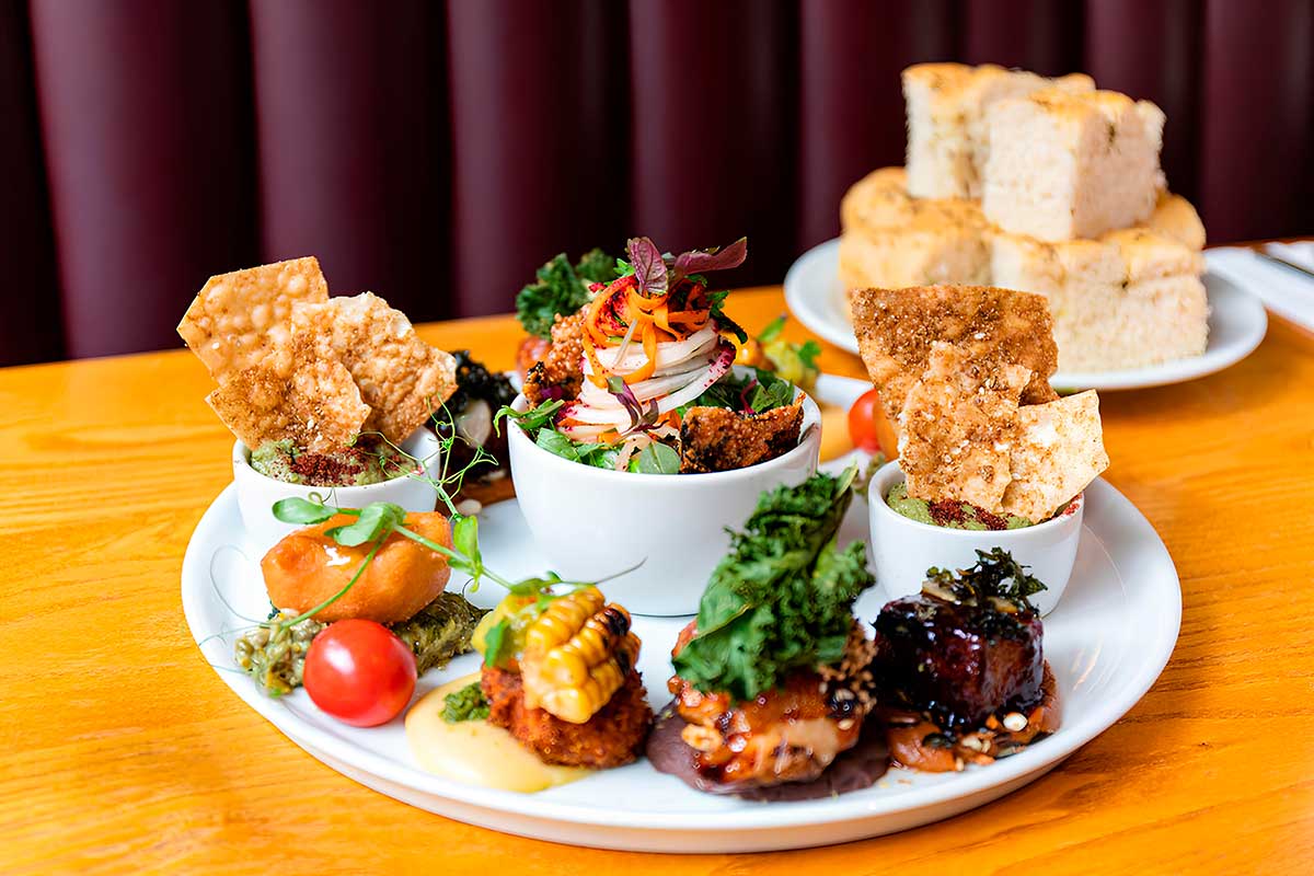 A vibrant vegan tapas platter from Terre à Terre in Brighton, featuring artfully presented small dishes with fresh vegetables, crisp crackers, sauces, and grilled corn, served with a side of focaccia bread.
