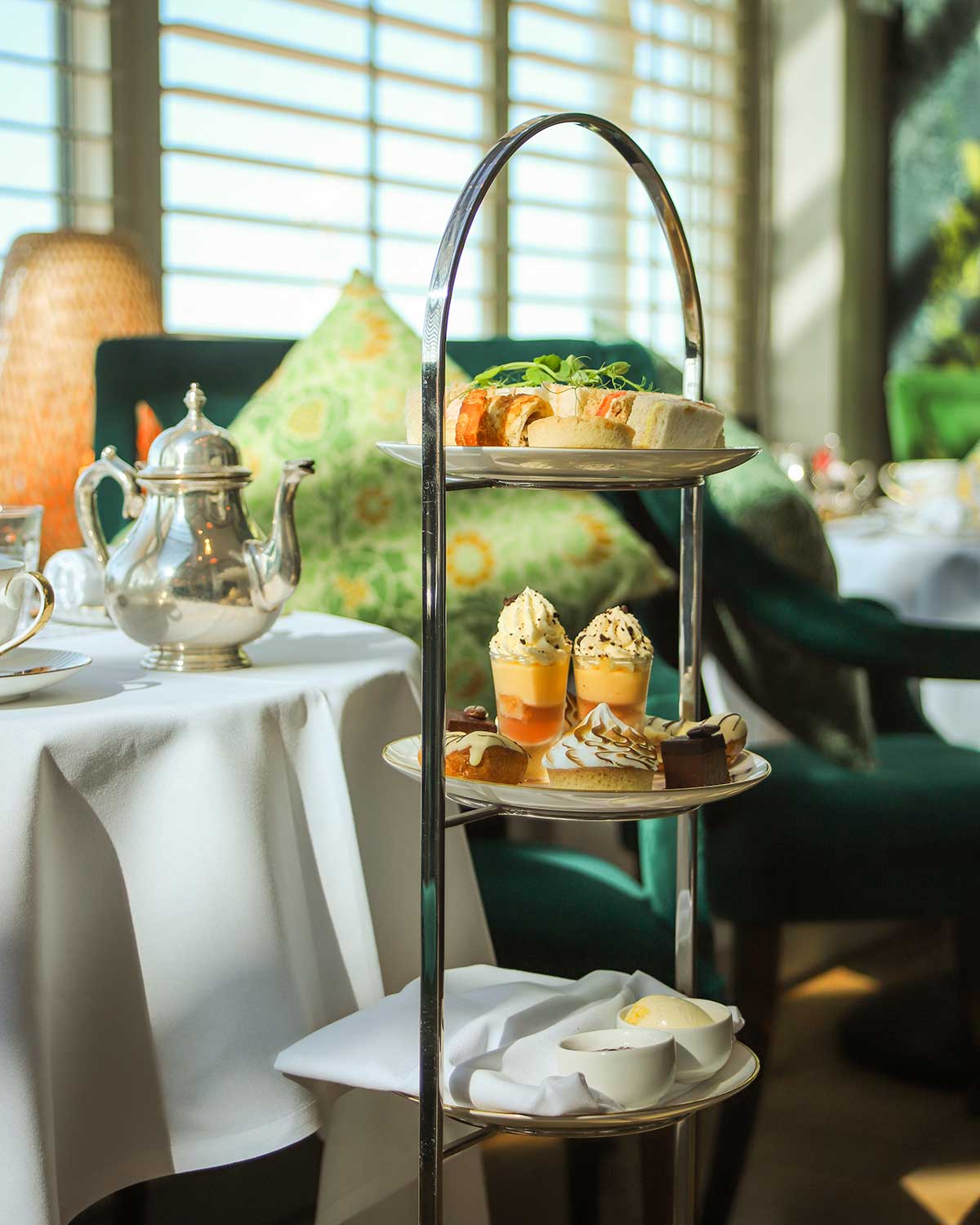 Elegant vegan afternoon tea at The Grand Brighton, featuring a silver tea stand with finger sandwiches and plant-based desserts, set beside a silver teapot on a white linen table in a sunlit lounge.