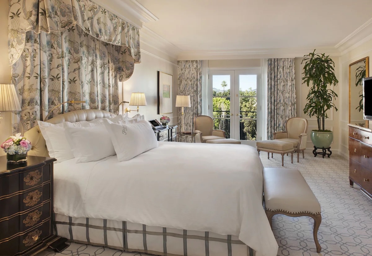 An elegant guest room at The Peninsula Beverly Hills vegan hotel in Los Angeles, featuring plush white bedding, floral drapes, and a balcony overlooking lush greenery.