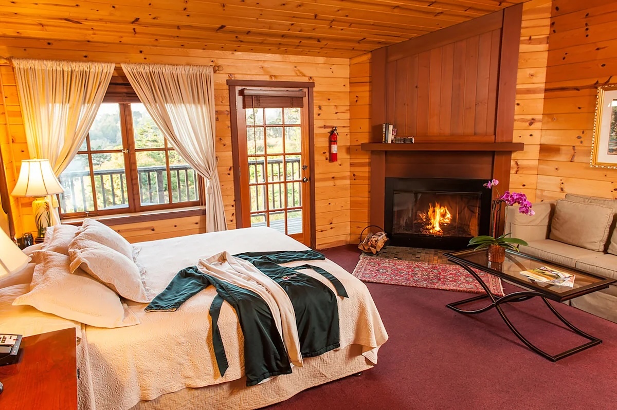 A cozy guest room at the vegan-friendly Stanford Inn by the Sea in Mendocino, California, featuring a wood-paneled interior, a wood-burning fireplace, a comfortable bed with plush white linens, and a balcony with views of lush greenery