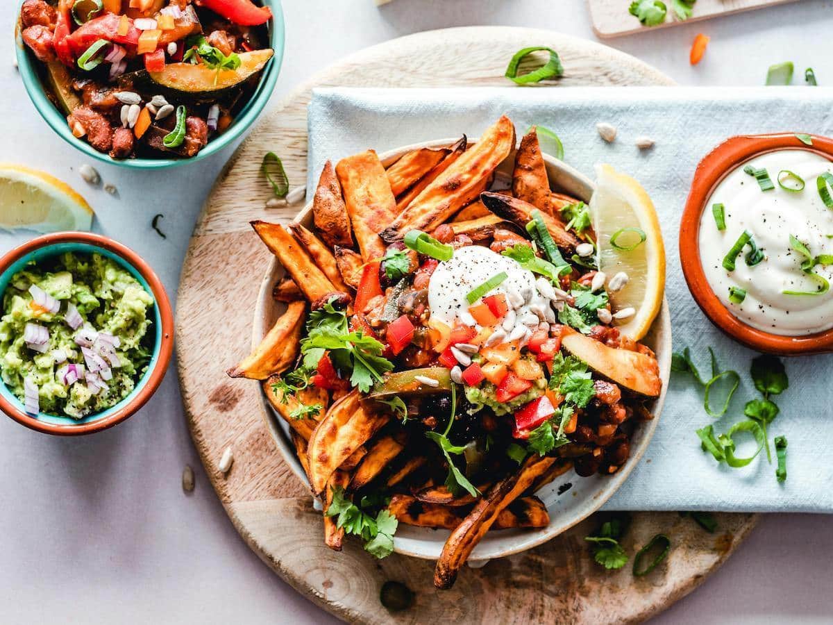 A vibrant vegan meal, with a main dish of sweet potato fries topped with fresh vegetables, herbs, sunflower seeds, and a dollop of vegan cream or yogurt. On the side, there are two small bowls, one containing guacamole with diced red onions and the other filled with a creamy dip garnished with chopped green onions. A lemon wedge is placed next to the dish for added zest.