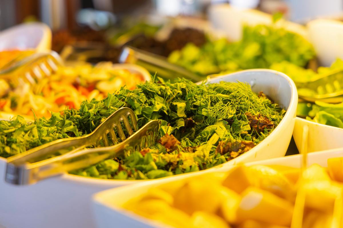 A vibrant salad bar at a vegan buffet. Large white bowls are filled with fresh greens, herbs, and vegetables. Metal tongs are placed in one of the bowls for easy serving, and other buffet options can be seen blurred in the background, showcasing a variety of healthy plant-based choices.