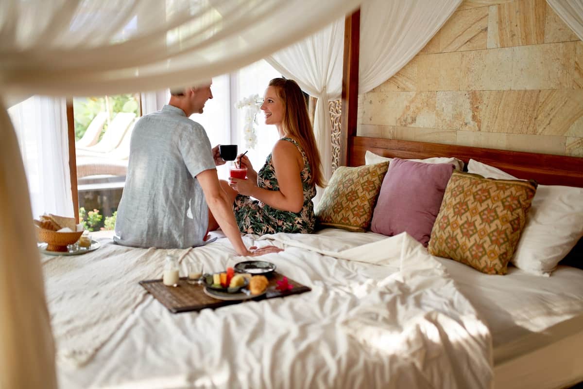 A couple enjoys a relaxed morning in a luxurious canopy bed with soft white linens at a tropical resort. They share coffee and fresh fruit while surrounded by elegant wooden decor, plush pillows, and an open-air view of a private pool and lush greenery.