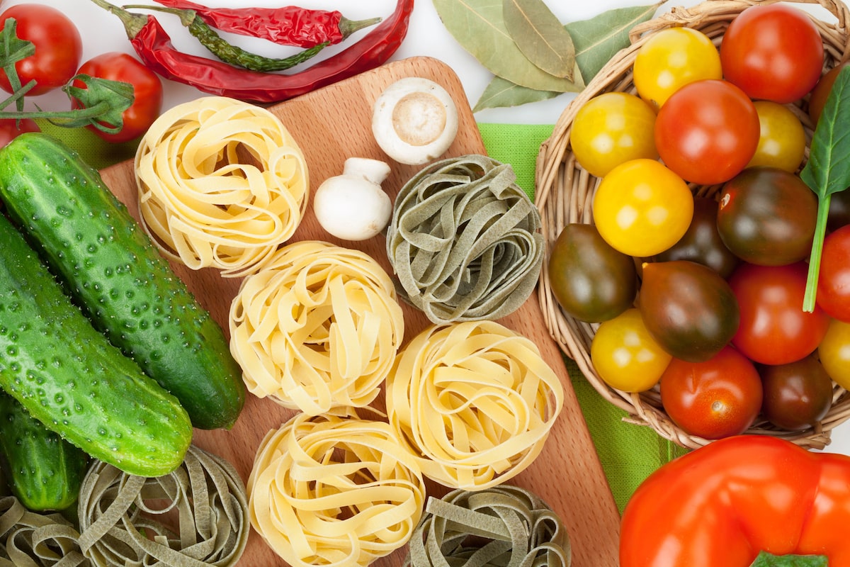 Flat lay of ingredients for vegan Italian cooking, including fresh cucumbers, cherry tomatoes, mushrooms, colorful tagliatelle nests, dried chili peppers, bay leaves, and bell pepper.