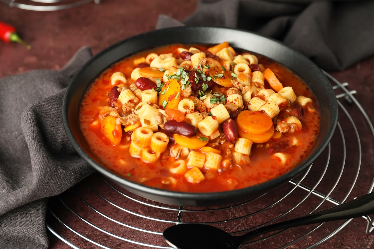 Bowl of vegan pasta e fagioli with ditalini pasta, kidney beans, carrots, and a tomato-based broth, garnished with herbs and served in a black bowl on a rustic background.