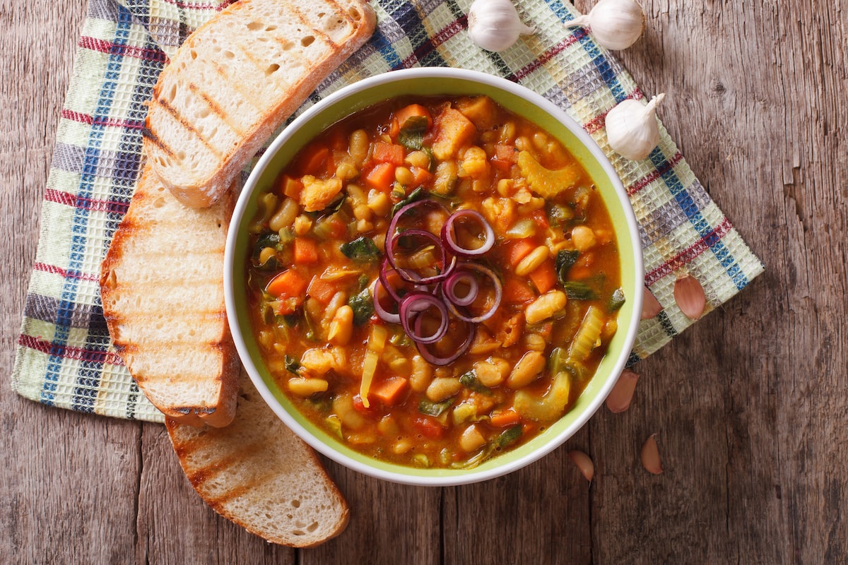 Bowl of vegan ribollita soup made with white beans, vegetables, and bread, served with grilled slices of Italian bread on a rustic wooden table.