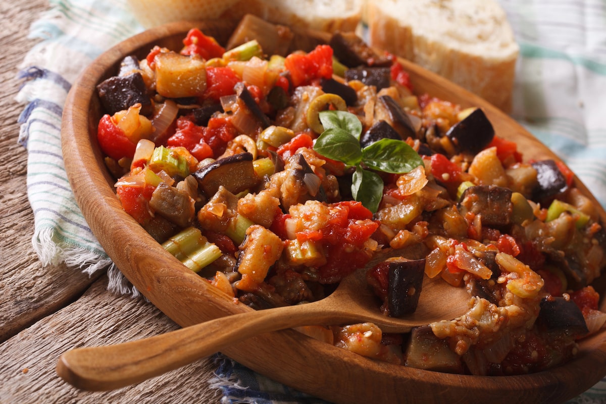 Caponata served in a wooden bowl with a spoon, featuring stewed eggplant, tomatoes, celery, and green olives, garnished with fresh basil.