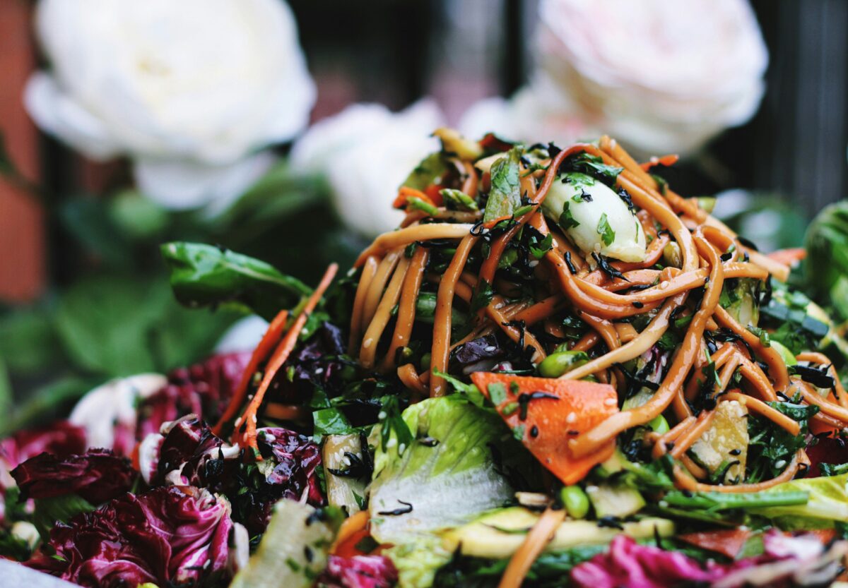 A tossed noodle dish with colorful vegetables, including carrots, leafy greens, and herbs, creating a fresh, textural mix. 
