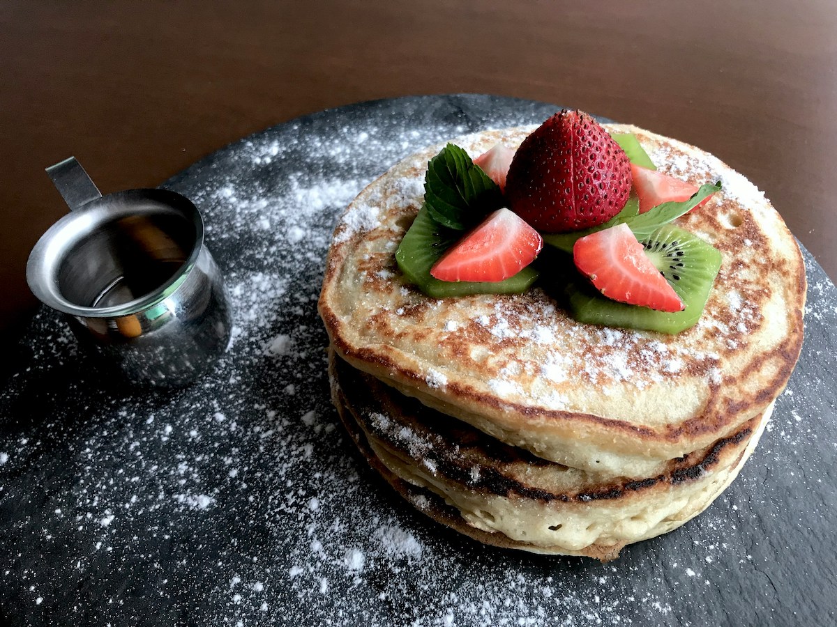 A stack of fluffy pancakes dusted with powdered sugar, topped with fresh slices of kiwi, strawberries, and a whole strawberry at the center. The pancakes are placed on a dark slate surface, with a small metal cup of syrup to the side.