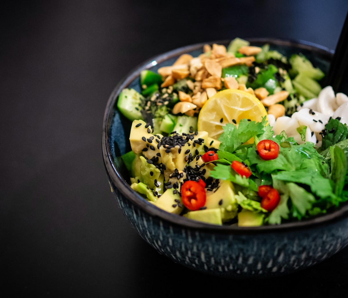 A vibrant bowl with a vegan salad. The salad features cubed avocado, cucumber, cilantro, slices of red chili peppers, sesame seeds, crushed peanuts, and a slice of lemon on top, all arranged in a black bowl with a dark background. 