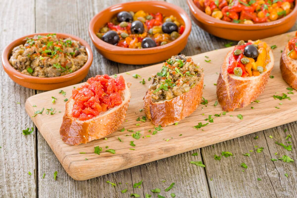 Assorted vegan Spanish tapas featuring a variety of vegetable toppings, including tomato bruschetta and an olive medley