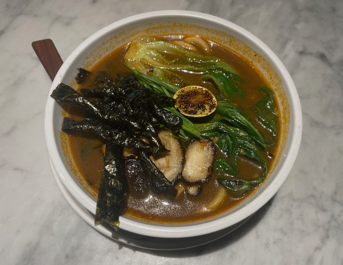 This image shows a bowl of vegan udon soup with rich broth, topped with fresh greens, nori strips, a slice of grilled lime, and shiitake mushrooms. The dish is presented in a white bowl on a marble surface, highlighting the vibrant colors of the ingredients.