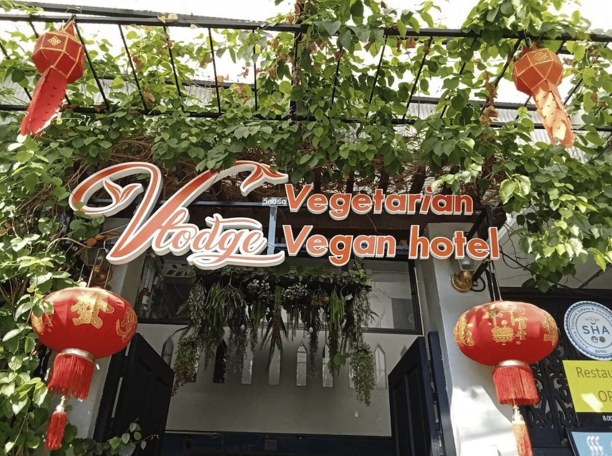 The entrance of Vlodge House, a vegan-friendly hotel located just outside of Chiang Mai's Old City in Thailand. The sign above the entrance reads 'Vlodge Vegetarian Vegan Hotel,' surrounded by lush green vines and traditional red lanterns, creating a welcoming and vibrant atmosphere.