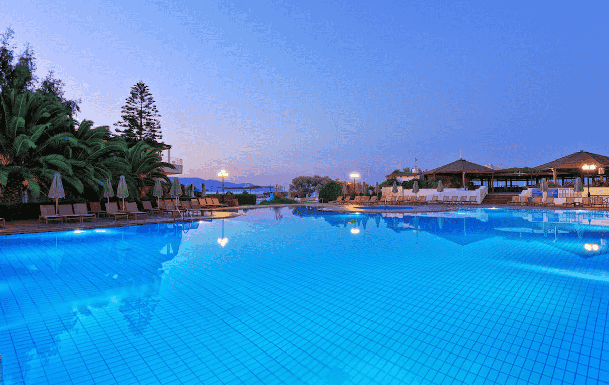 A breathtaking twilight view of the expansive pool at Apollonia Beach Resort & Spa, surrounded by palm trees, sun loungers, and softly glowing lights. The resort’s open-air pavilions and serene coastal backdrop create a tranquil and luxurious atmosphere for guests.