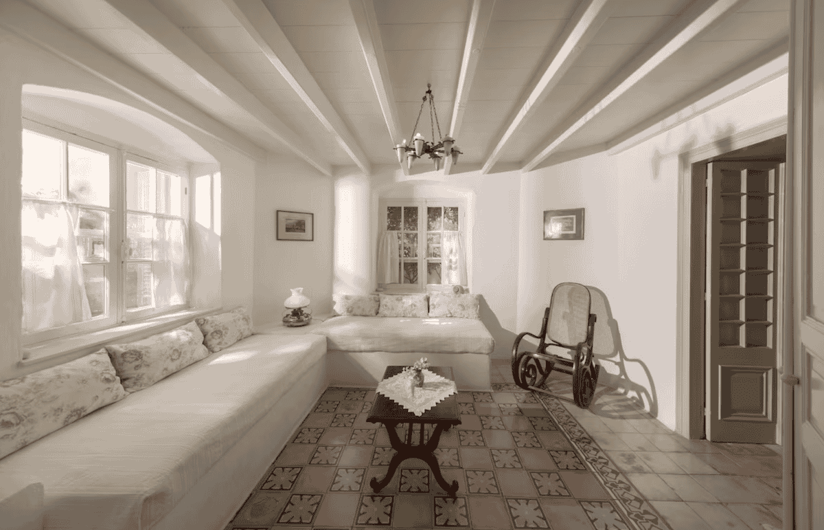 A charming vintage-style living room at Archontiko Angelou, a historic boutique hotel in Greece. The space features white wooden beams, elegant built-in sofas with floral cushions, a classic rocking chair, and a small wooden coffee table over decorative tiled flooring, evoking a nostalgic and cozy atmosphere.