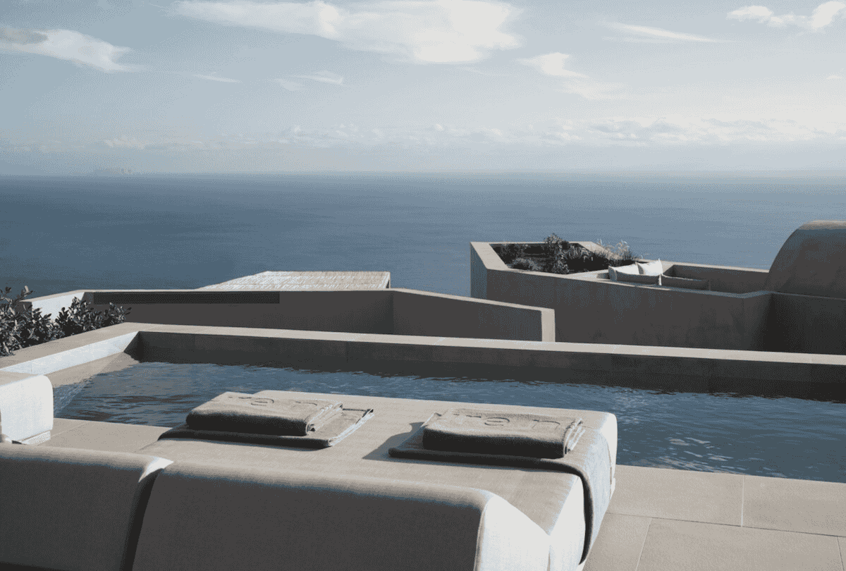A luxurious infinity pool at Ethos Vegan Retreat in Imerovigli, Santorini, overlooking the vast Aegean Sea. The modern, minimalist terrace features sun loungers with neatly folded towels, blending seamlessly with the tranquil, earthy architecture.