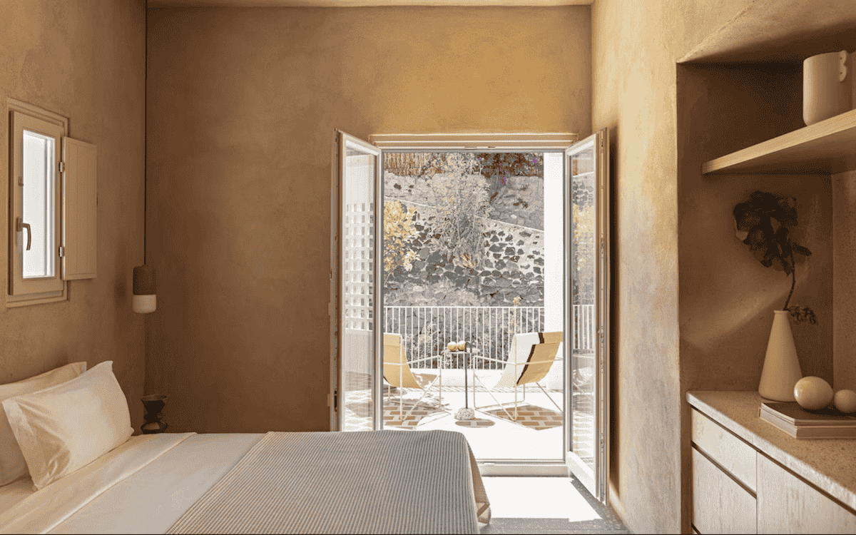 A serene bedroom at Ethos Vegan Suites in Greece, featuring earth-toned walls, minimalist decor, and an open balcony with lounge chairs overlooking a stone wall garden.