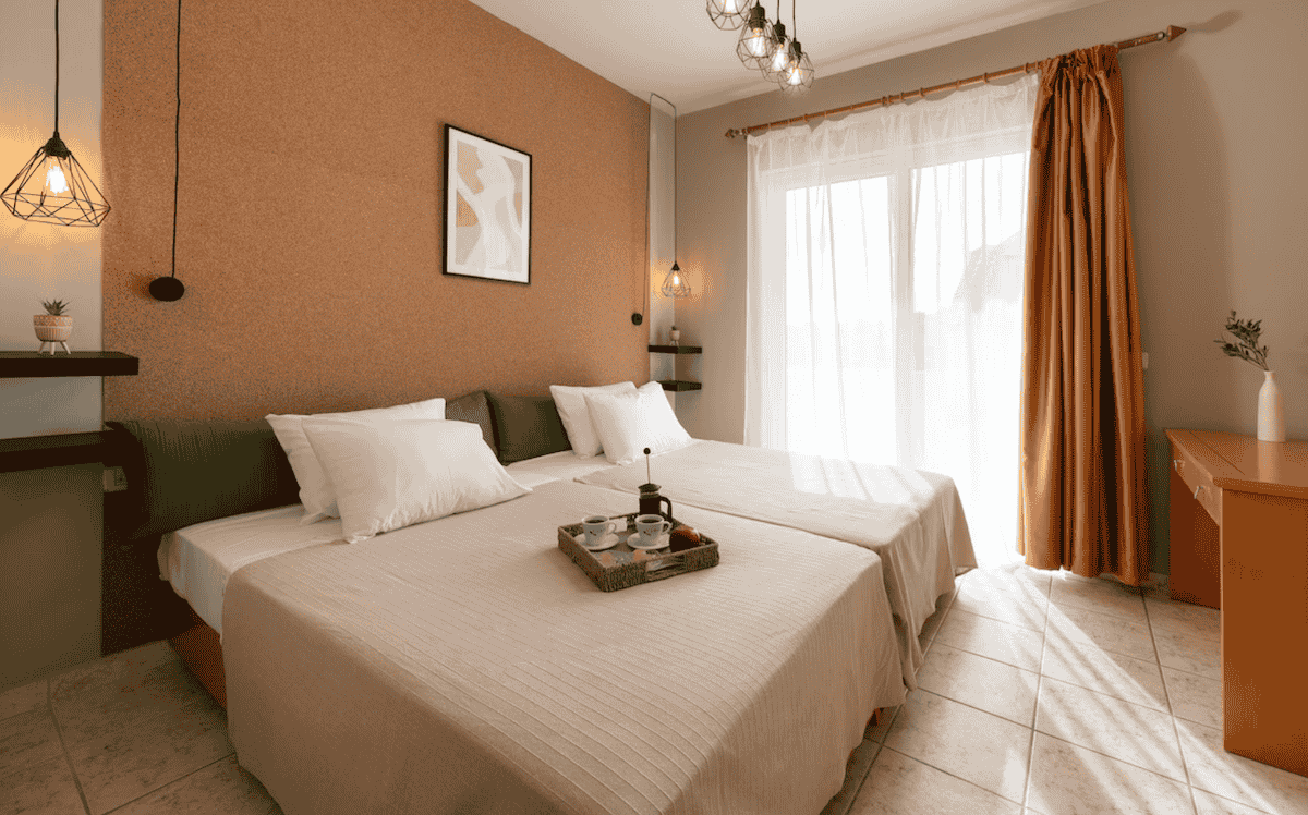 A cozy and stylish twin bedroom at GOJI Vegan Hotel, featuring earthy tones, warm lighting, and modern pendant lamps. A breakfast tray with coffee and pastries sits on the neatly made beds, while sheer curtains allow soft natural light to fill the inviting space.