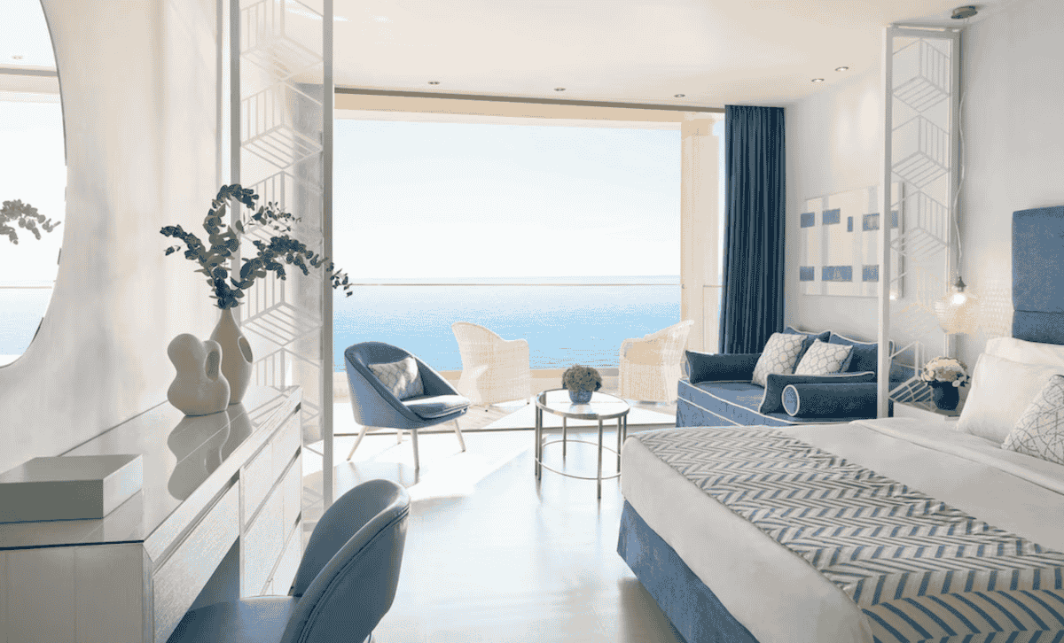 A bright and elegant oceanfront suite at Ikos Oceania, featuring a blue and white color scheme inspired by the Aegean Sea. The room includes a plush bed, stylish seating area, and a private balcony with panoramic sea views, creating a luxurious and serene retreat.